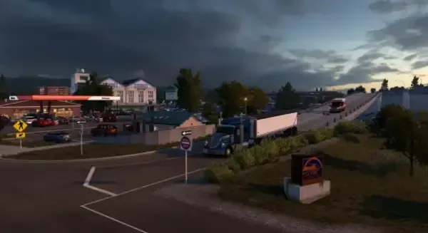 A truck driving through a small town at dusk, with a gas station and various buildings in the background. The sky is overcast, and there are trees and signs along the roadside. 