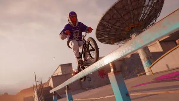 A BMX rider in a purple hoodie and white pants performing a grind on a blue rail at sunset, with a satellite dish in the background. The scene is set in an urban skate park with graffiti-covered buildings and a clear sky.