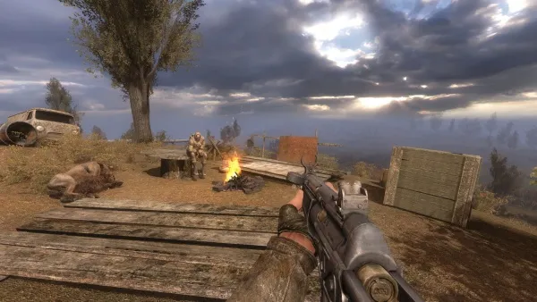 A first-person perspective of a video game scene set in a post-apocalyptic world. The player is holding a rifle with a scope, aiming at two figures standing near a campfire. There's a dilapidated car and a large tree nearby. The sky is overcast with rays of sunlight breaking through the clouds. The environment appears desolate and abandoned.