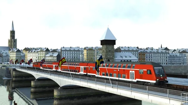 A red train crossing a bridge over a river, with European flags on the bridge and historic buildings in the background. The scene is set during winter, as indicated by the snow-covered roofs and ground. The architecture includes a prominent church tower and a fortified gatehouse. The sky is clear with soft sunlight casting shadows.